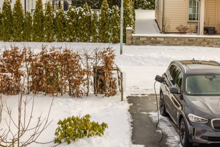 Téléchargez les photos : Vue du parking dans villa privée avec voiture électrique reliée par câble depuis la station de recharge le jour d'hiver. Suède. - en image libre de droit