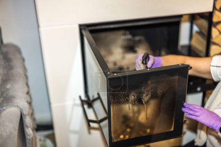 View of woman's hand using a spray to clean soot from the glass door of a fireplace. Sweden.