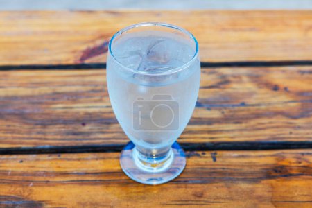 Nahaufnahme eines mit Eiswürfeln gefüllten Glases, das mit Kondenswasser bedeckt und mit kaltem Wasser auf einem Holztisch in einem Restaurant gefüllt ist.