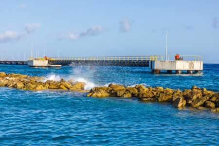 Blick auf das Karibische Meer mit einem Steg und einem künstlichen Wellenbrecher, an dem Wellen gegen die Felsen krachen. Curaçao.