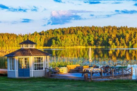 Beautiful view of a lakeside gazebo and deck with outdoor furniture, surrounded by lush trees and their reflections on the calm water. Sweden.