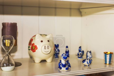 Close-up view of shelf display featuring decorative piggy bank with floral designs, an hourglass, candle, and various small blue and white ceramic figurines.