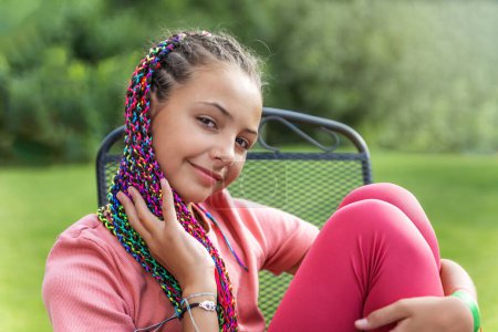 Side view of young girl with colorful braids in her hair sitting on the metal garden chair outside. Horizontally. 