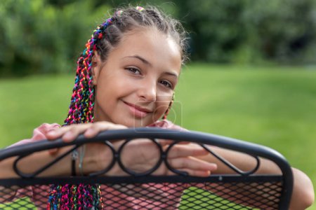 Nahaufnahme Vorderseite Porträt eines jungen Mädchens mit bunten Zöpfen im Haar sitzt auf dem Metallstuhl draußen. Horizontal. 