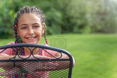Lächelndes und glückliches junges Mädchen mit bunten Zöpfen im Haar, das draußen auf dem Stuhl sitzt. Horizontal. 