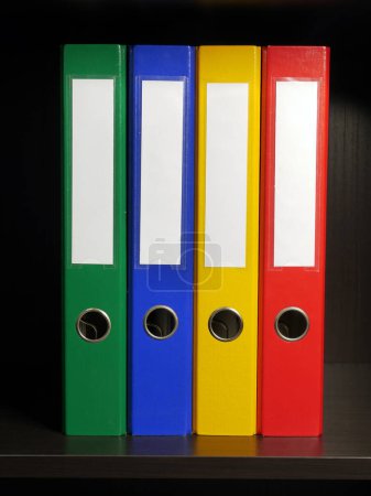 Téléchargez les photos : Quatre classeurs vides en vert, bleu, jaune et rouge disposés à l'intérieur de l'armoire noire - en image libre de droit