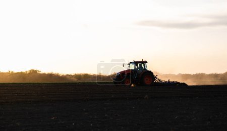 Un agriculteur dans un tracteur prépare son champ alors que le soleil commence à se coucher. 
