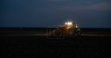 Tractor preparing land with seedbed cultivator at night.