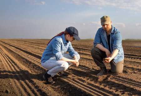 Foto de Jóvenes agricultores examinando la tierra mientras el tractor está arando el campo - Imagen libre de derechos