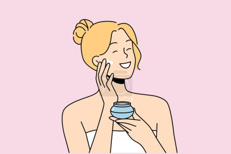 Woman applies anti-aging cream to face to stay beautiful, standing in bath towel after shower. Happy blonde girl smiles and uses cosmetic cream to help prevent wrinkles and pimples.