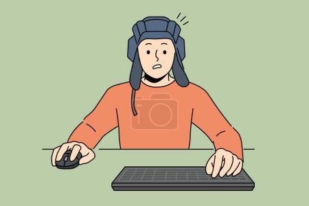 Man gamer plays tanks on computer, sitting at table with keyboard and wears tanker helmet. Guy gamer passionate about video games and virtual tank simulators, makes embarrassed grimace after losing
