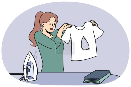 Ungeschickte Hausfrau bügelt Kleidungsstücke im T-Shirt und weiß nicht, wie sie Fehler beheben soll. Frau steht mit Kleidung und Bügeleisen neben Bügelbrett und betrachtet beschädigten Gegenstand verwirrt.