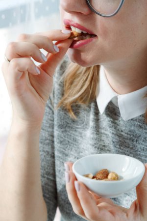 Woman Eating Nuts, une collation saine pleine de minéraux. Gros plan de bouche femelle mordant l'écrou et tenant la plaque blanche avec la hanche de différentes noix