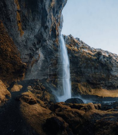 Foto de Cascada de Kvernufoss en la garganta de las montañas. Atracción Turística Islandia. Hombre con chaqueta roja de pie y mira el flujo de agua que cae. Belleza en la naturaleza
. - Imagen libre de derechos