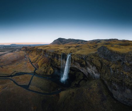 Téléchargez les photos : La prise de vue aérienne de Seljalandsfoss est située dans la région sud de l'Islande. Les visiteurs peuvent marcher derrière. Cascade Seljalandsfoss avec un grand coucher de soleil sur la destination touristique populaire. Une partie de l'or - en image libre de droit