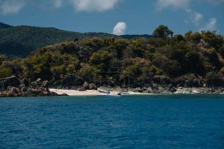 Téléchargez les photos : Un bateau flotte paisiblement dans les eaux cristallines près d'une île verte luxuriante, entourée d'un paysage naturel pittoresque de montagnes et de végétation - en image libre de droit