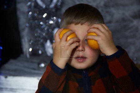 Téléchargez les photos : Adorable garçon joyeux tout-petit joue avec les mandarines parmi les décorations de Noël. Maison atmosphérique festive pour le réveillon du Nouvel An. Esthétique maison confortable. Joyeux petit garçon. Joyeux concept. Photo de haute qualité - en image libre de droit