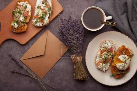Table with sandwiches of Greek yogurt and zucchini spread, poached egg, salmon, coffee cup, envelope and bouquet of lavender top view