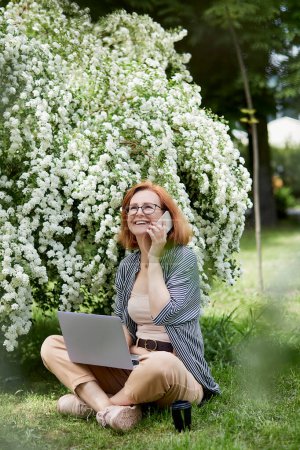 Photo for Happy young woman speaking by phone, using laptop in white flowering blooms. Remote work, chatting with friends, studying concept. - Royalty Free Image