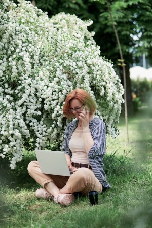 Photo for Young woman with red hair is chatting on her phone while working on her laptop outdoors, surrounded by white blossoms. - Royalty Free Image