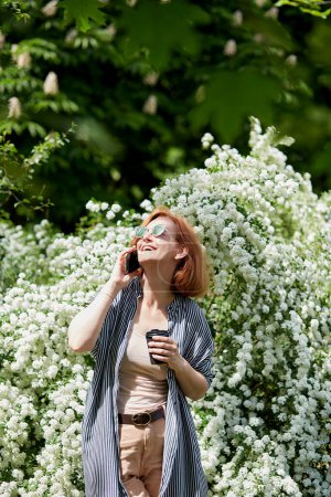 Photo for Red haired woman speaks by phone. Nature's call in freelance life a joyful work moment under the blooming branches - Royalty Free Image