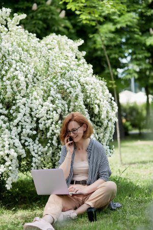 Happy young woman speaking by phone, using laptop in white flowering blooms. Chatting with friends, remote work, studying concept.