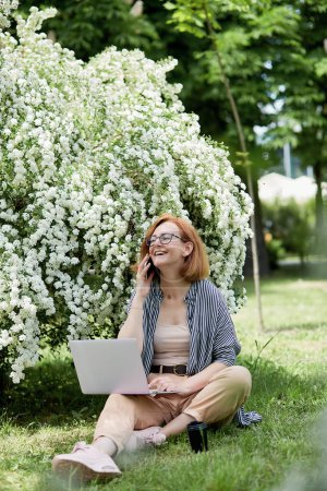 Glückliche junge Frau lächelt mit Handy und Laptop in weiß blühenden Blüten. Fernarbeit, Chat mit Freunden, Konzeptstudium.