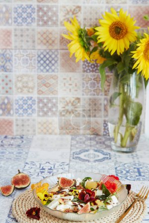 Vegetarischer Salat mit Feta, essbaren Blumen, Feigen, Pfirsich, Basilikum auf Keramikfliesen Hintergrund mit Sonnenblumen. Coze Hausküche.