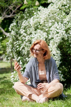 Young smiling red-haired woman using her phone, seated outdoors by white flowering blooms with coffee cup.