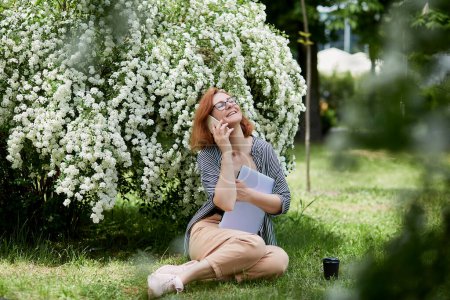 Soleado día de trabajo disfruta de un freelancer disfruta de una conversación en un parque floral. Mujer pelirroja habla por teléfono con el ordenador portátil.