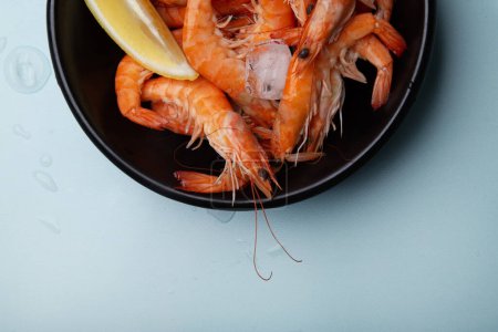 Bowl full of defrosted cooked prawns with lemon wedges, on a blue background with droplets of water. Ideal for culinary websites or seafood recipes.