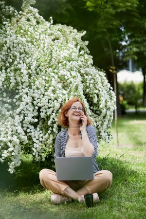 Photo for Happy young woman speaking by phone, using laptop in white flowering blooms. Remote work, chatting with friends, studying concept. - Royalty Free Image