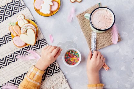 Delightful Easter, child adorns cookies with colorful sprinkles flat lay.