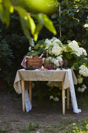Panier de pique-nique d'été avec couverture, bouteille vintage, fruits frais et verres violets, contre des arbustes d'hortensias. Représentation tranquille des loisirs et du mode de vie gastronomique