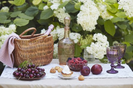 picnic basket with blanket, vintage bottle, fresh fruits, and purple glasses, set against hydrangea bushes