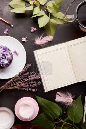 Estilo de vida estético femenino. Burla de cuaderno abierto entre taza de café, magdalena floral púrpura, crema facial y flores.