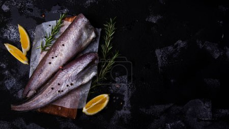 Fresh raw fish hake garnished with rosemary and lemon slices on a dark textured surface, perfect for culinary themes or healthy eating concepts. Extra wide banner