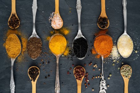 Many different exotic spices on tablespoons of metal and small wooden spoons arranged in a row in horizontal, landscape format on a large slate plate and additional peppercorns