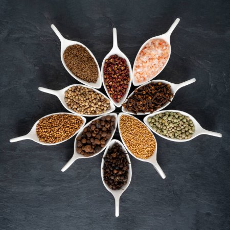 Exotic spices from all over the world in small white porcelain bowls arranged in a circle on a large black slate plate