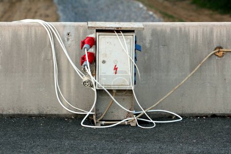 Photo for Heavy industrial outdoor metal electrical high voltage box with multiple outdoor electrical outlets and safety switches connected with thick electrical cables mounted on concrete wall near paved road at local construction site on warm summer day - Royalty Free Image