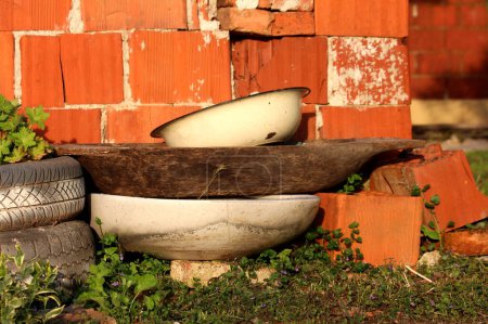 Téléchargez les photos : Bunch of old junk in suburban family house backyard next to red building blocks wall consisting of old white partially rousted metallic washbowl on top of rustic wooden bread trough or pulgh trough or kneading trough or artesa - en image libre de droit