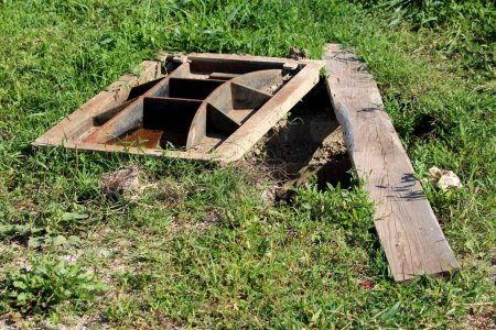 Large heavy custom made rectangle cast metal partially rusted manhole cover next to open manhole temporarily covered with dilapidated wooden board surrounded with uncut grass at local field on warm sunny summer day