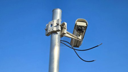 Newly installed large white old style closed circuit TV CCTV surveillance camera mounted on strong grey metal pole with large metal mount waiting to be connected with two loose black electrical wires on clear blue sky background