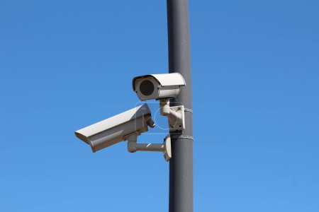 Two small fake dummy modern white closed circuit TV CCTV security cameras looking in opposite directions mounted on dark grey metal pole on clear blue sky background at local construction site
