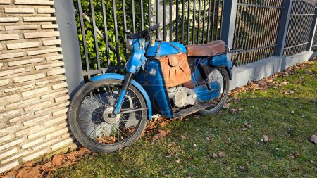 Vintage old in need of restoration blue motorbike with worn out light brown leather seat next to rustic handmade leather motorcycle saddle bag and small decorative lantern keychain chained to grey modern metal fence of suburban family house