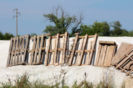 Reihenweise kaputte alte Holzpaletten auf einer provisorischen Hochwasserschutzwand aus Kastenbarrieren und Sandsäcken, die mit dickem, weißem Geotextilgewebe überzogen sind, das Familienhäuser vor massiven Überschwemmungen bei starkem Regen schützen soll 