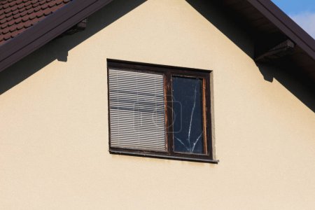 Téléchargez les photos : Fenêtre mansardée avec cadre en bois fissuré délabré et stores fermés sur la gauche et verre cassé patché avec ruban transparent sur le côté droit sur le dessus de la maison familiale urbaine le jour chaud ensoleillé d'automne - en image libre de droit