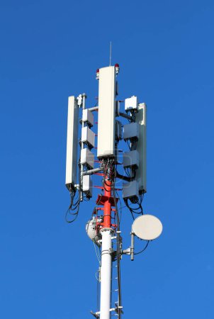 Verschiedene Formen und Größen Handysender und Antennen, die auf einem hohen, starken roten und weißen Metallmast auf klarem, blauem Himmelshintergrund montiert sind