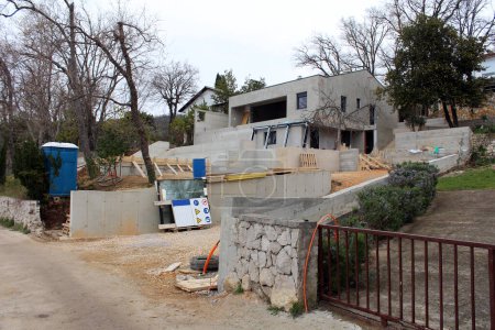 Construction site for modern villa by the sea filled with construction material mixed with wooden supports and glass windows waiting to be installed surrounded with trees without leaves and portable unisex blue with white top ecological toilet