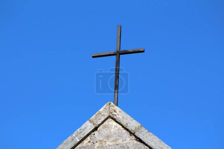 Dilapidated old worn down black metal cross on top of traditional Catholic stone church partially covered with yellow moss on clear blue sky background